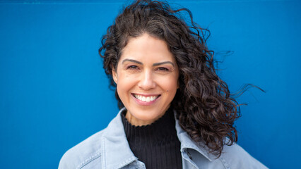 Confident woman with curly hair smiling at the camera against a bright blue background, wearing a...