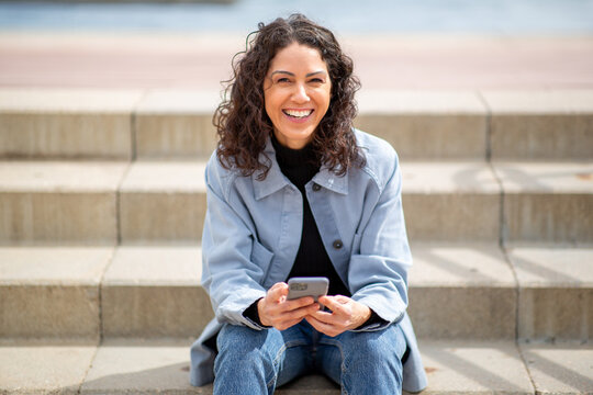 Cheerful woman with curly hair sitting on outdoor steps, smiling at the camera while holding a smartphone