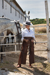 A young, beautiful Spanish woman dressed in riding clothes and wearing a traditional Spanish hat stands next to a horse in the countryside on a sunny day. More horses graze in the background.