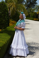 A beautiful young Spanish woman dressed in a traditional white Andalusian flamenco dress with a flower in her hair. The woman picks up a yellow flower with her hand and poses for the photo.