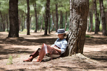 A young, pretty woman dressed in riding breeches and riding clothes and a typical Spanish hat rests seated in the shade of a pine tree on a pilgrimage day in Spain.