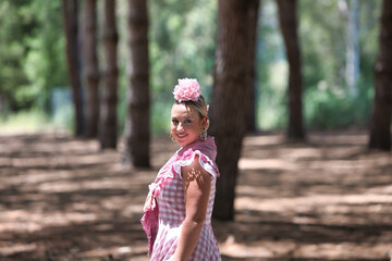 A beautiful young Spanish woman dressed in a traditional pink and white flamenco dress with a flower in her hair walks among the pine trees of Andalusia during the pilgrimage.