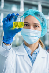 An scientist woman wearing laboratory mask and protective gloves, holding a small beaker filled with liquid while working in modern laboratory. Scientific research concept