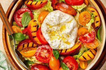 Close up of burrata cheese on a summer salad with grilled peaches, tomatoes, black pepper and dressed with olive oil. Top view.