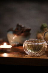 Transparent glass cup filled with tea placed on wooden tea tray. Small burning tea light candle creating peaceful, cozy atmosphere. Tea ceremony aesthetics with feelings of comfort and warmth 