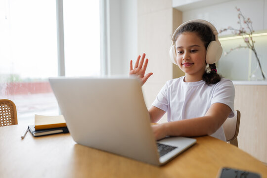 Girl engaging in a virtual learning session at home while wearing headphones and smiling warmly during a video call with classmates