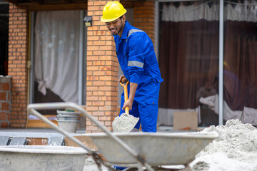 Construction worker shoveling sand at building site. Construction worker wearing a blue jumpsuit and yellow helmet is shoveling sand at a building site, working on a new residential project.