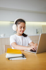 Young girl engaged in online studying while wearing headphones in a bright, modern kitchen during the day