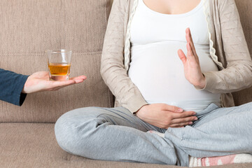 Close up of pregnant woman sitting on the sofa refuses to drink whisky and makes stop gesture to...