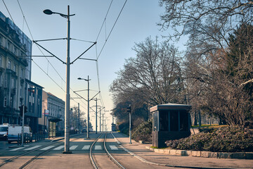 Early morning view of quiet urban street with empty tram tracks running down the center. Overhead power lines and street infrastructure frame the composition. Melancholic european city atmosphere