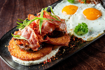 Delicious breakfast with sunny-side-up eggs, crispy hash browns, grilled bacon, and fresh herbs, served on a dark plate with rustic wooden
