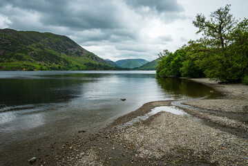 A moody landscape unfolds from the pebble beach of Ullswater, where calm lake waters meet distant...