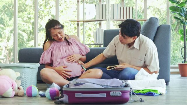 Pregnant wife experiences sudden labor pains while packing her clothes and belongings while her husband watches over her with concern and calls the hospital for emergency assistance.