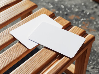 Two blank white rectangular cards with rounded corners, casually placed on a slatted wooden bench in sunlight.
