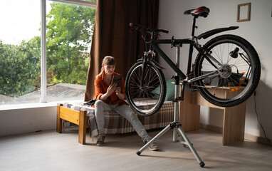Middle aged woman repairing bicycle sitting on bed using digital tablet to search for spare parts...