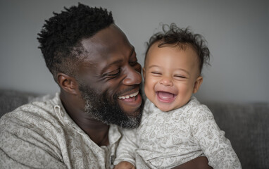 A Black father wearing a white t-shirt with his baby son sitting on the bed, smiling at each other in the bedroom at home. African American dad and child laughing together while playing indoors.