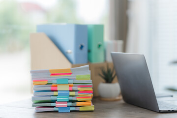 Work Desk Essentials: A stack of documents marked with colorful sticky notes sits next to a sleek laptop, with office supplies in the background, all under the soft, natural light of a window.