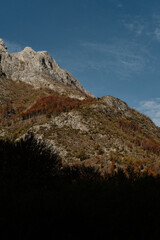 Sharp ridges and colorful forested slopes of the Prokletije mountains in autumn, captured in warm light under a clear blue sky. Nature of Montenegro.