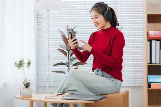 Woman in Video Call: An Asian woman, adorned in a vibrant red sweater and wearing headphones, engrossed in a video call via her smartphone, radiating happiness and a connection with technology.