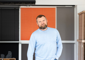 A small business owner stands confidently in his sunshade systems showroom. Surrounded by roller blinds, pleated shades, and wooden blinds, he proudly showcases his custom window covering solutions.