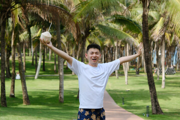 cheerful Asian young man raising coconut joyfully in palm trees grove in Sanya, Hainan, China