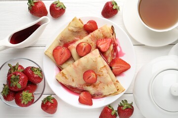 Delicious crepes with strawberries, sauce and tea on white wooden table, flat lay