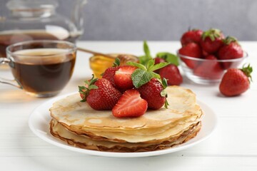Delicious crepes with strawberries, honey, mint and tea on white wooden table, closeup