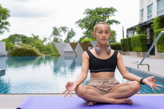 Genderfluid individual with mustache and lipstick relaxing poolside while wearing a bikini-style top. Confident, expressive LGBTQ+ representation in summer lifestyle setting.