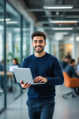 A young man with dark hair and a beard confidently smiles while holding a laptop in a modern office setting.