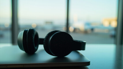 Black headphones resting on a closed silver laptop near a window with a blurred airport background during daytime