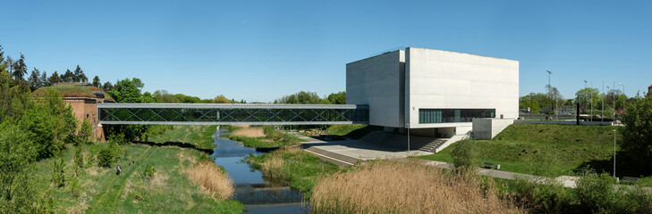 Obraz premium Brama Poznania museum blends modern glass architecture with historical brickwork, set by a river and lush greenery, symbolizing cultural harmony.