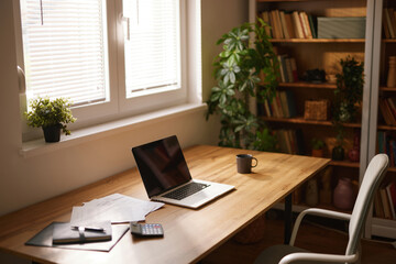 Fototapeta premium A neatly arranged home office includes a laptop, documents, and a coffee cup on a wooden desk, illuminated by natural light from a nearby window and surrounded by plants.
