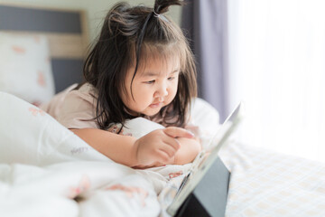 A little Asian girl is lying on the bed watching a tablet,Asian girl lying on bed in bedroom and...