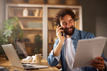 A man with a beard holds papers in one hand and talks on the phone with a smile. He sits at a wooden desk, surrounded by books and plants, in a well-lit home office.