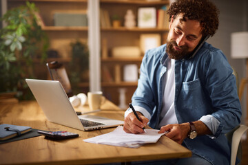 A man sits at his home office desk engaged in a phone conversation while jotting down notes. A...