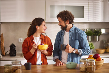 In a bright and modern kitchen, a couple happily shares breakfast. The woman holds a bowl of...