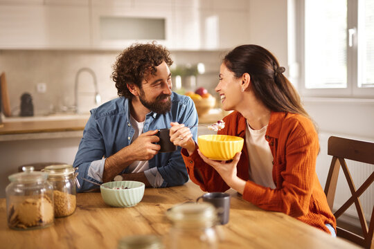 A couple sits at a wooden table in a well-lit home office, sharing a light breakfast with smiles, enjoying each other's company and a moment of connection.
