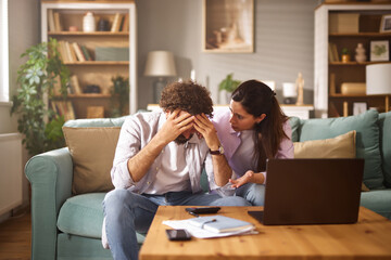 A man is visibly stressed, holding his head in his hands, while a woman seated next to him points...