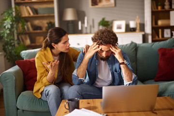 A woman expresses concern to a man sitting on a couch, who appears stressed while looking at a...