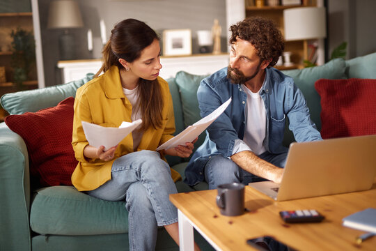 A couple engages in a serious discussion about their financial issues while sitting together on a couch, surrounded by documents, a laptop, and a coffee mug at their home.