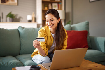 A woman smiles while holding a credit card and using her laptop at home. She is relaxed on a couch,...
