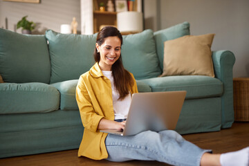 A woman sits on the floor in a casual outfit, focused on her laptop in a comfortable living room...