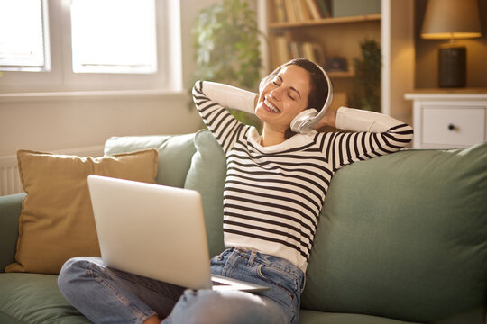 A young woman smiles in a relaxed position, enjoying music on a comfortable couch in a bright living room. Light filters through the windows, creating a warm atmosphere.