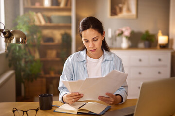 A woman sits at a wooden desk in a softly lit home office, examining financial papers. She appears...
