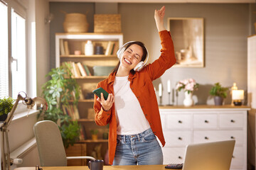 A woman wearing headphones smiles and dances while holding a smartphone in her stylish and...
