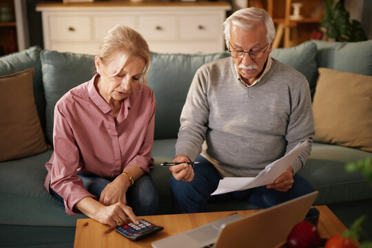 An older man and woman sit on a sofa at home, examining paperwork and using a calculator and laptop. They appear to be managing their finances or discussing insurance options.