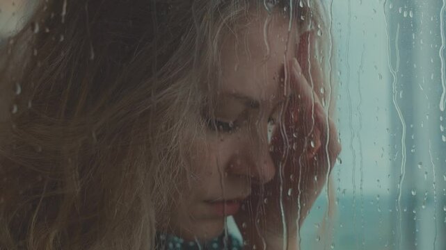 young adult unhappy woman crying standing at window rain drops water drip flow down the glass, depression sadness emotion. sad tears grief face eyes. scared lonely lady in panic attack hands shaking
