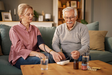 A senior woman sits on a couch and takes her blood pressure using an electronic monitor while her...