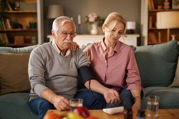 An elderly man checks his blood pressure with an automatic monitor while sitting on a couch. His...