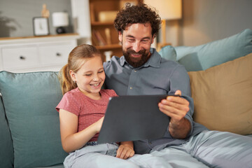 A father and his daughter are sitting on a couch, smiling and watching a movie together on a tablet...